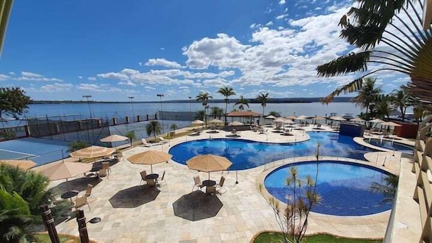 A resort pool area with multiple pools, lounge chairs, umbrellas, and palm trees. - Luxuoso Flat em Reort com vista incrível (Brasília)