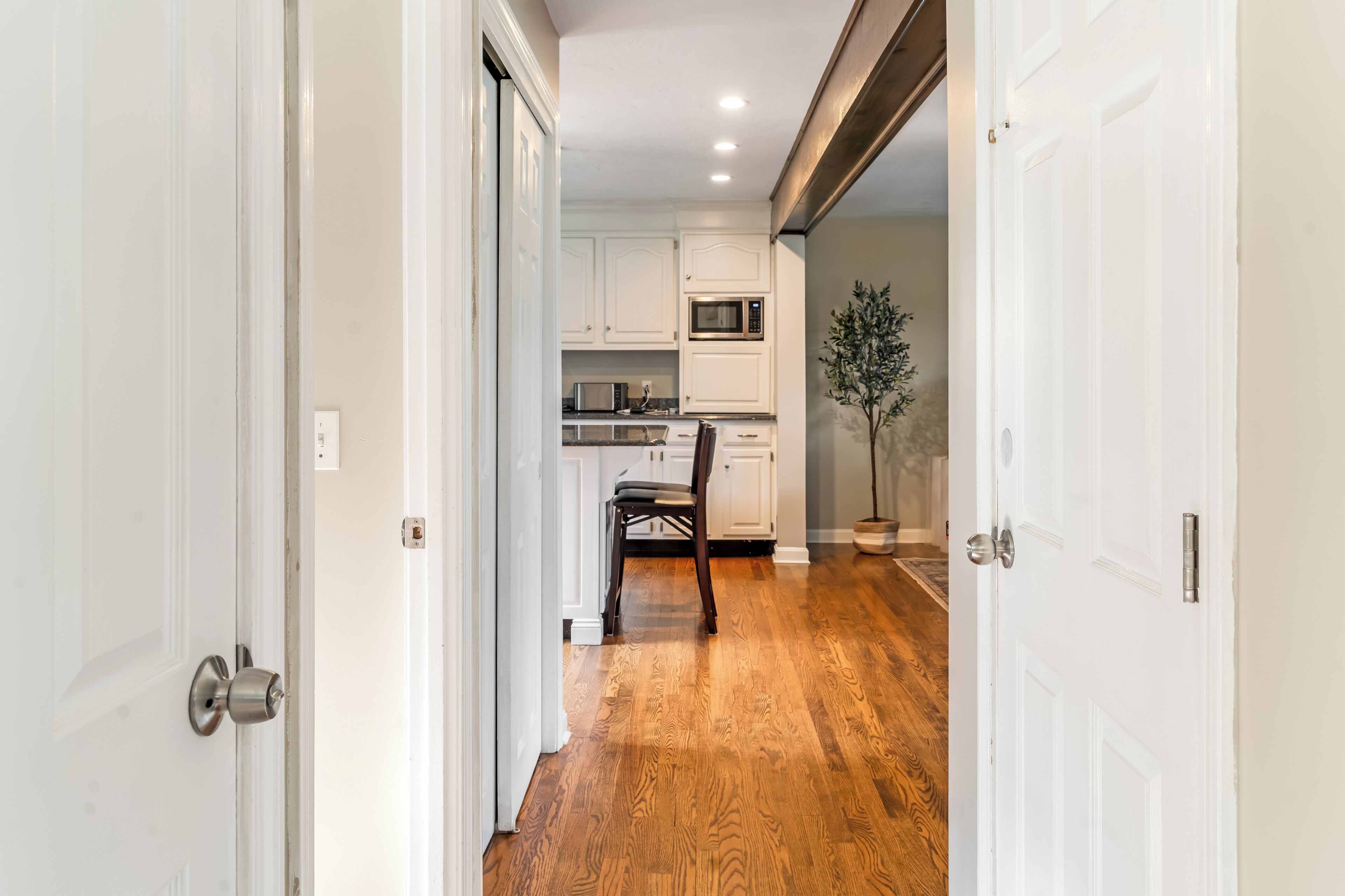 A hallway leading to a kitchen with white cabinets, a microwave, and a dining area with a chair.