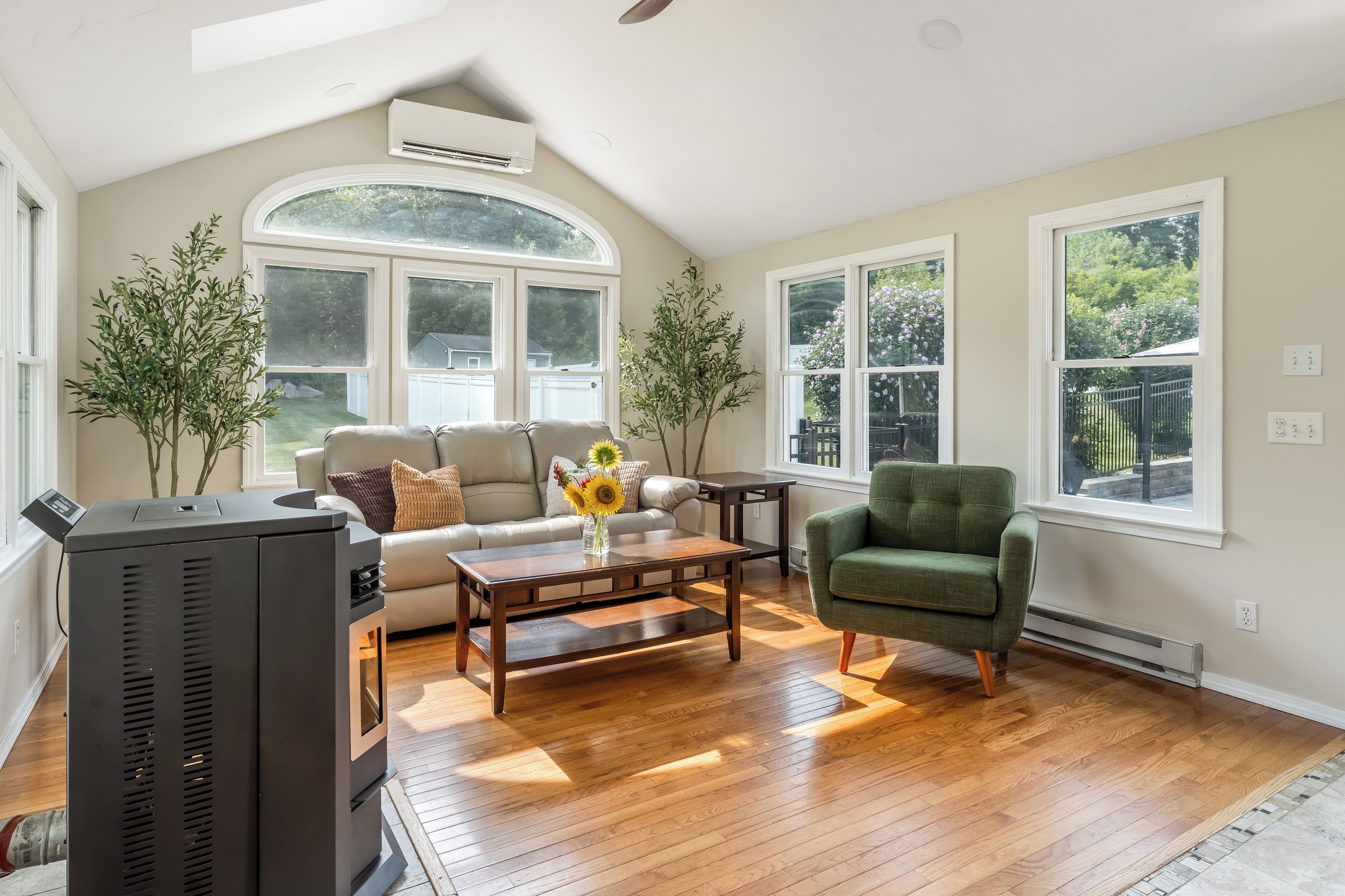 A living room with a sofa, armchair, coffee table, and a fireplace. There are large windows and a view of the outdoors.