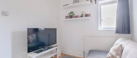 Un salon avec un téléviseur à écran plat posé sur un meuble blanc, un canapé gris, un radiateur blanc et une fenêtre avec un rideau.