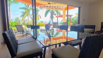 Comedor moderno con mesas de vidrio y sillas de mimbre, con vista a una piscina y palmeras.
