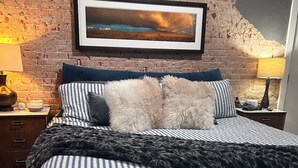 Een slaapkamer met een bakstenen muur, een lijst met een landschapsafbeelding, een bed met gestreepte beddengoed, zachte kussens, een donkere plaid, een nachtkastje met een lamp, en een klein decoratief object.