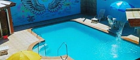 Una piscina con un mural de alas y una fuente, rodeada de sillas de descanso y un paraguas amarillo.