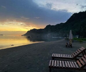 A beach with wooden lounge chairs and a single umbrella, surrounded by a forested cliff. - Bayview Cabin Retreat (Sipalay, Negros Occidental)