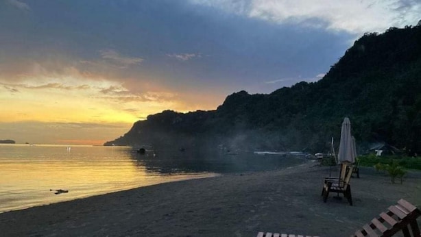 Una playa con sillones de madera y un paraguas, rodeada de un acantilado cubierto de vegetación.