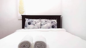 Una cama bien hecha con toallas enrolladas al frente, un cabecero oscuro y una pared con un cojín estampado con hojas.