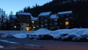 A multi-story building with a snow-covered roof, surrounded by parked cars and a snow-covered ground.