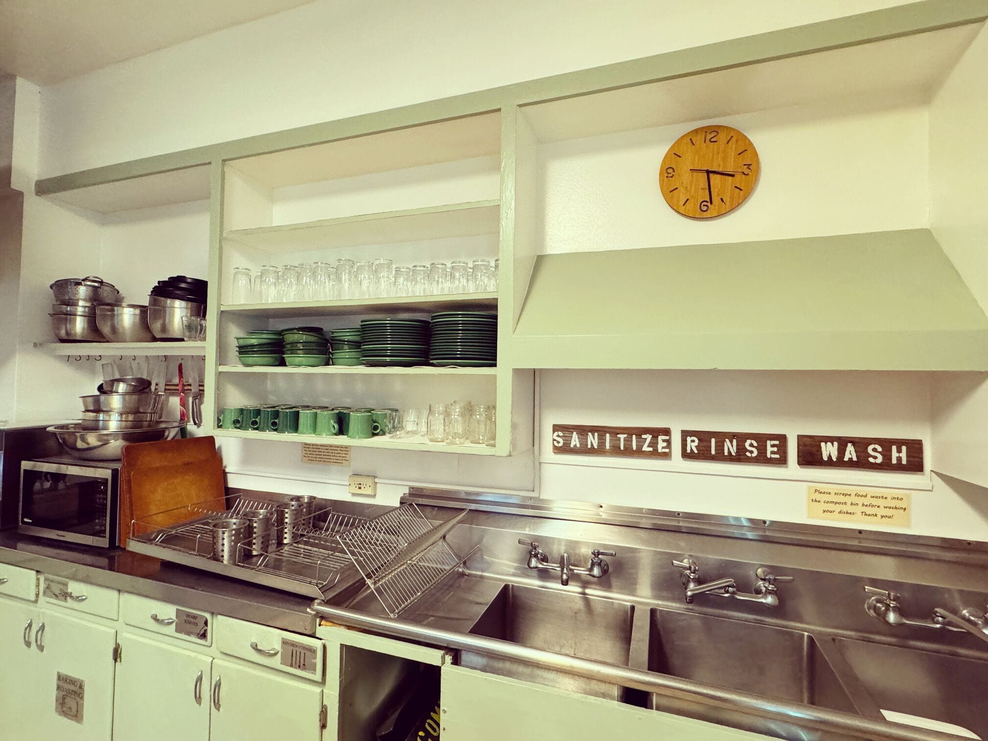 A kitchen with stainless steel sinks, shelves with dishes, pots, and a clock.