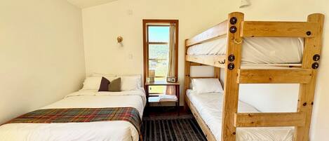 A room with a single bed, a bunk bed, a small table with a radio, and a window with a view of greenery.