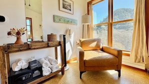 A room with a large window, a wooden console table with towels, a chair, and a mirror.