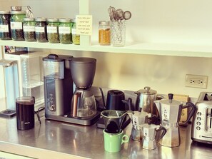 A kitchen counter with various coffee-making equipment, including a Krups coffee maker, a French press, and a moka pot. There are also several jars of coffee beans and a sign requesting labels on stored food.