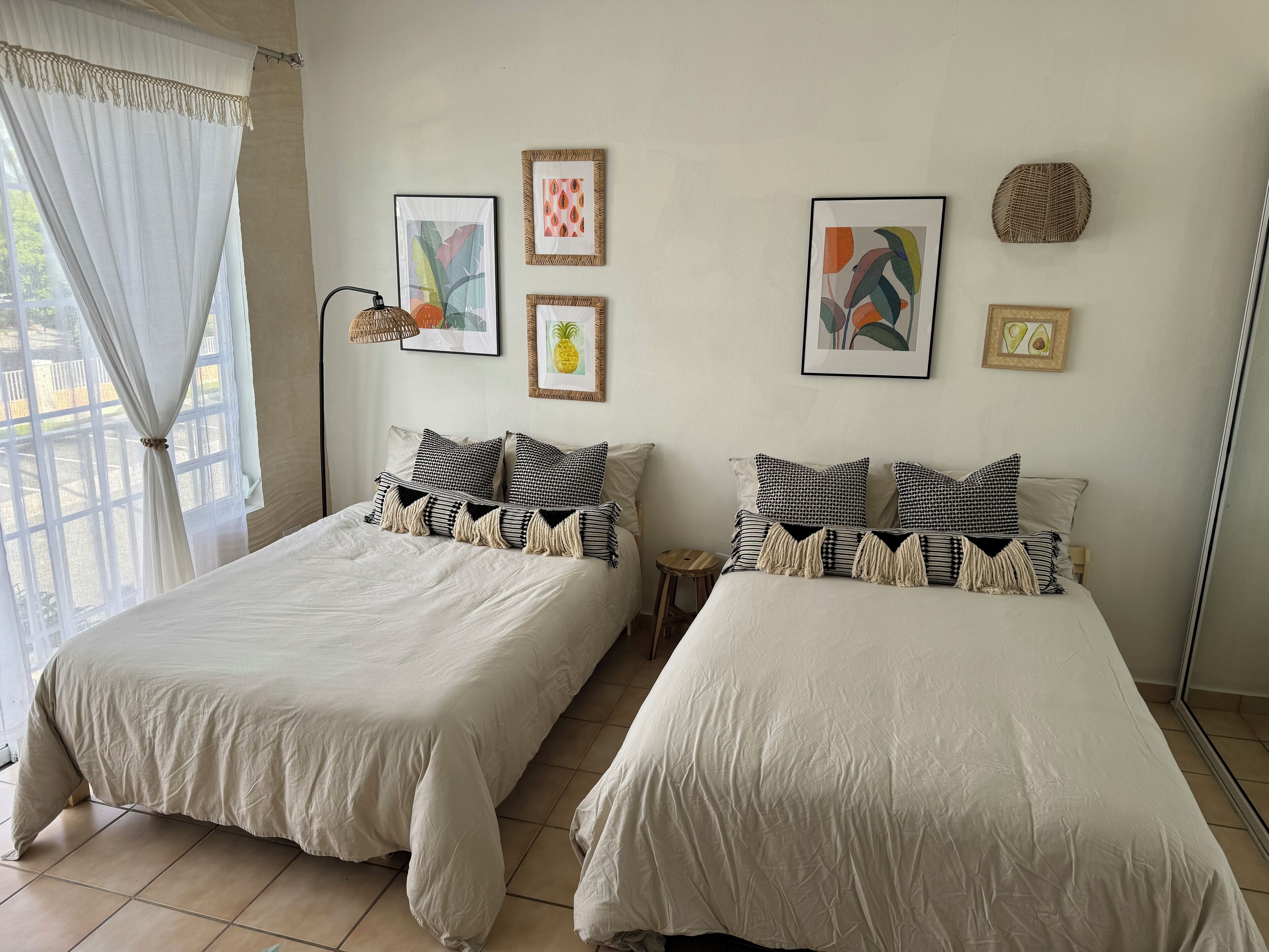 Duas camas com roupa de cama e almofadas iguais, uma janela com cortinas leves e uma obra de arte na parede.