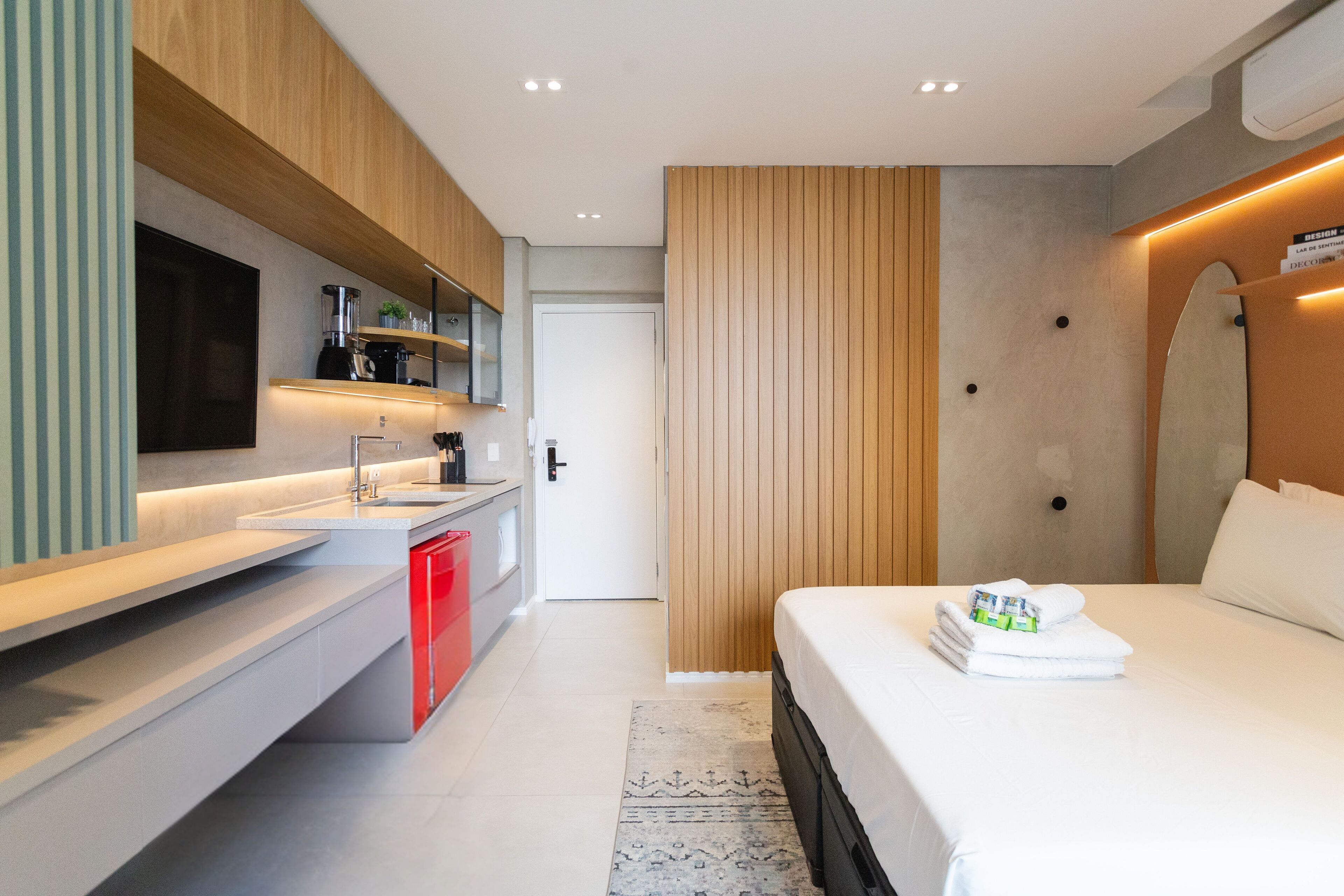 Ein modernes Hotelzimmer mit Bett, Nachttisch, integriertem Fernseher und Küchenzeile inklusive Spüle und Schränken.