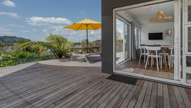Una terraza de madera con un paraguas amarillo, un área de comedor con sillas blancas y un televisor en la pared.
