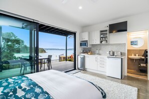 Ett modernt sovrum med en stor säng, en balkong med utsikt över havet och ett kök med vita skåp och en platt-TV.
