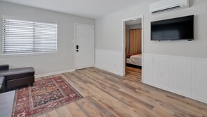 Ein Zimmer mit Holzboden, einem großen Fenster mit Jalousien, einer Tür, einem an der Wand befestigten Flachbildfernseher und einem kleinen Teppich.