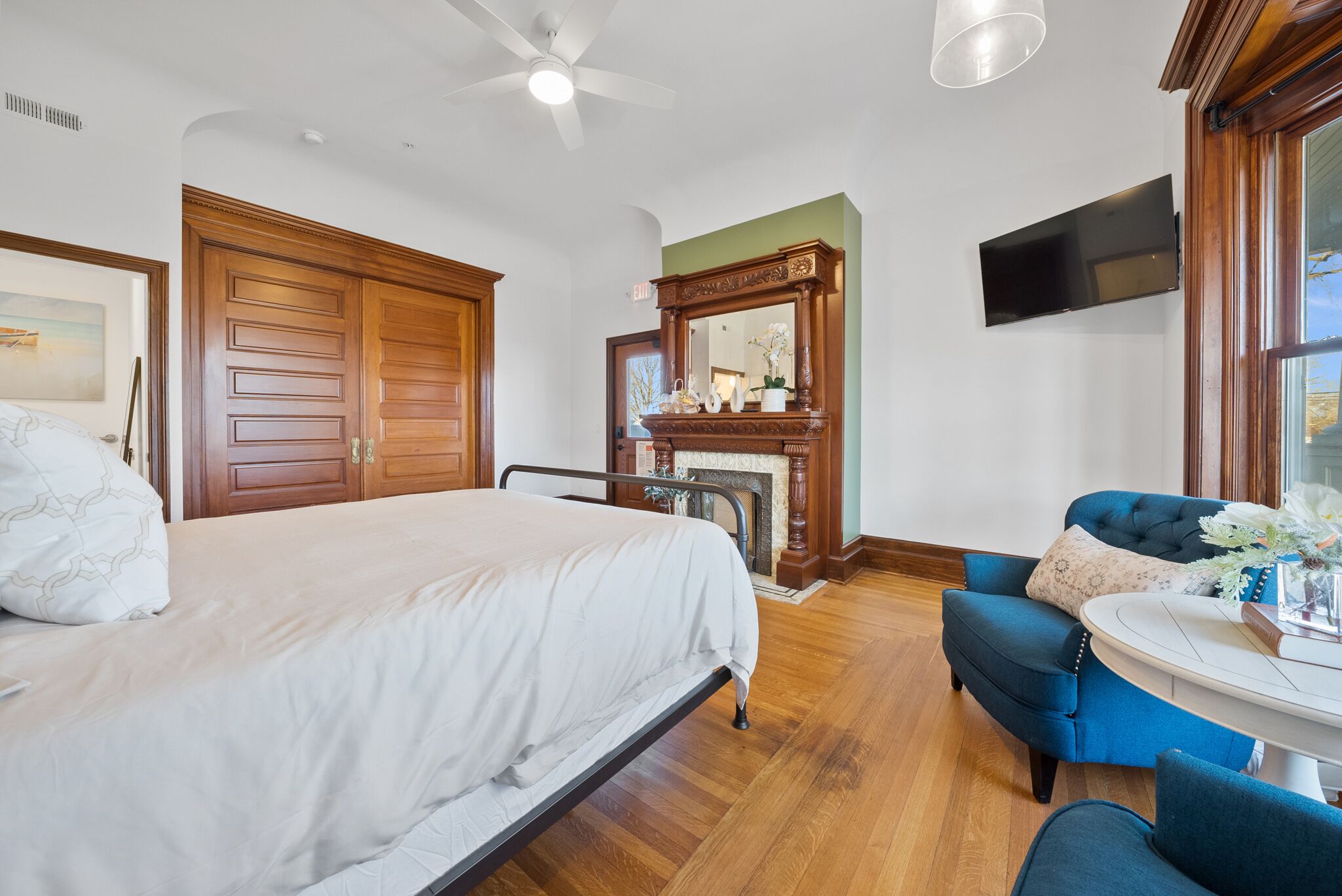 A bedroom with a bed, a blue armchair, a wooden wardrobe, a fireplace, and a television mounted on the wall.