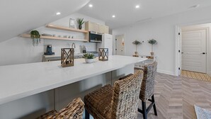 Cozinha moderna com armários brancos, bancada branca e micro-ondas embutido. Há duas cadeiras de vime e um pequeno vaso com planta no balcão.