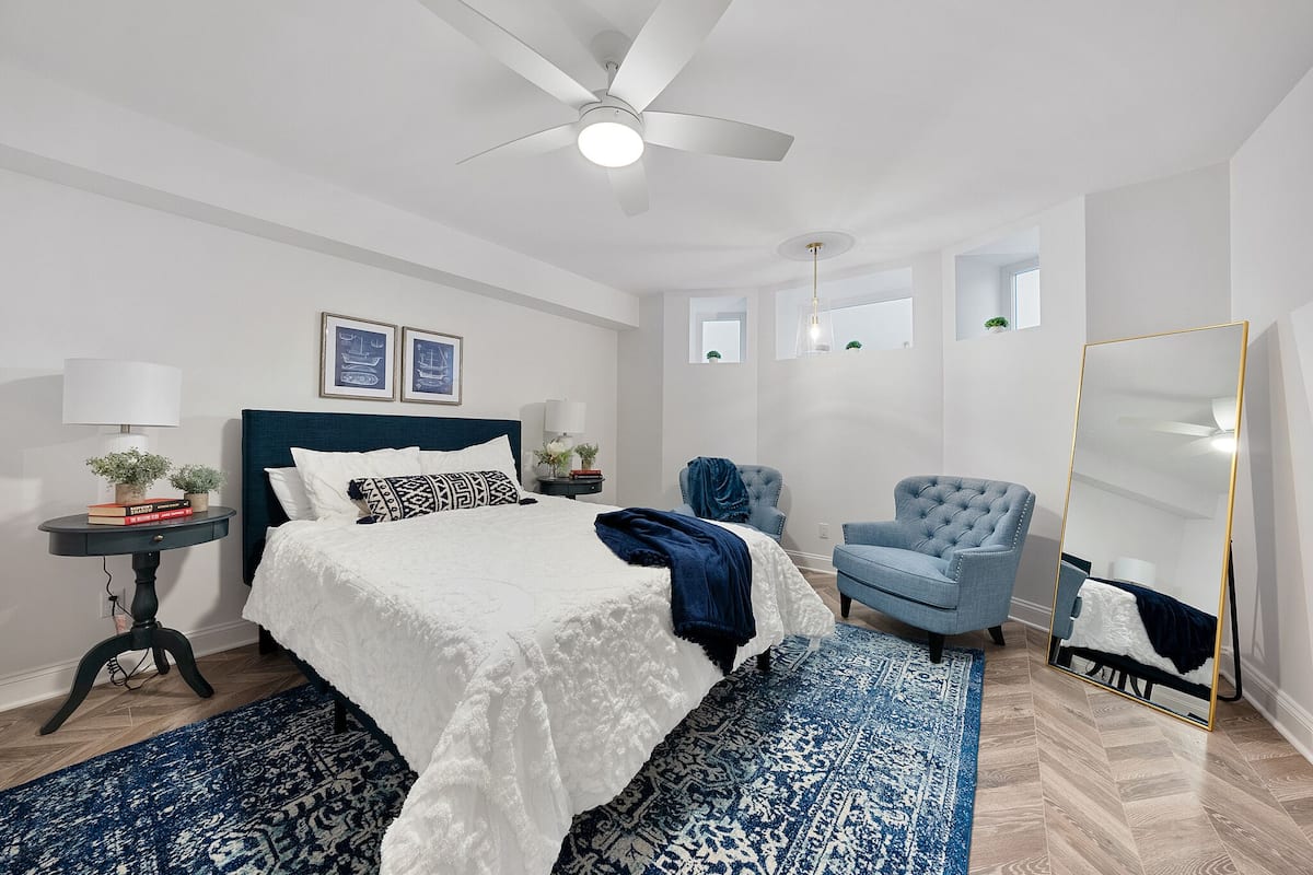 Un dormitorio con una cama, un ventilador de pie, un sillón azul, un espejo y una mesita de noche con una lámpara.