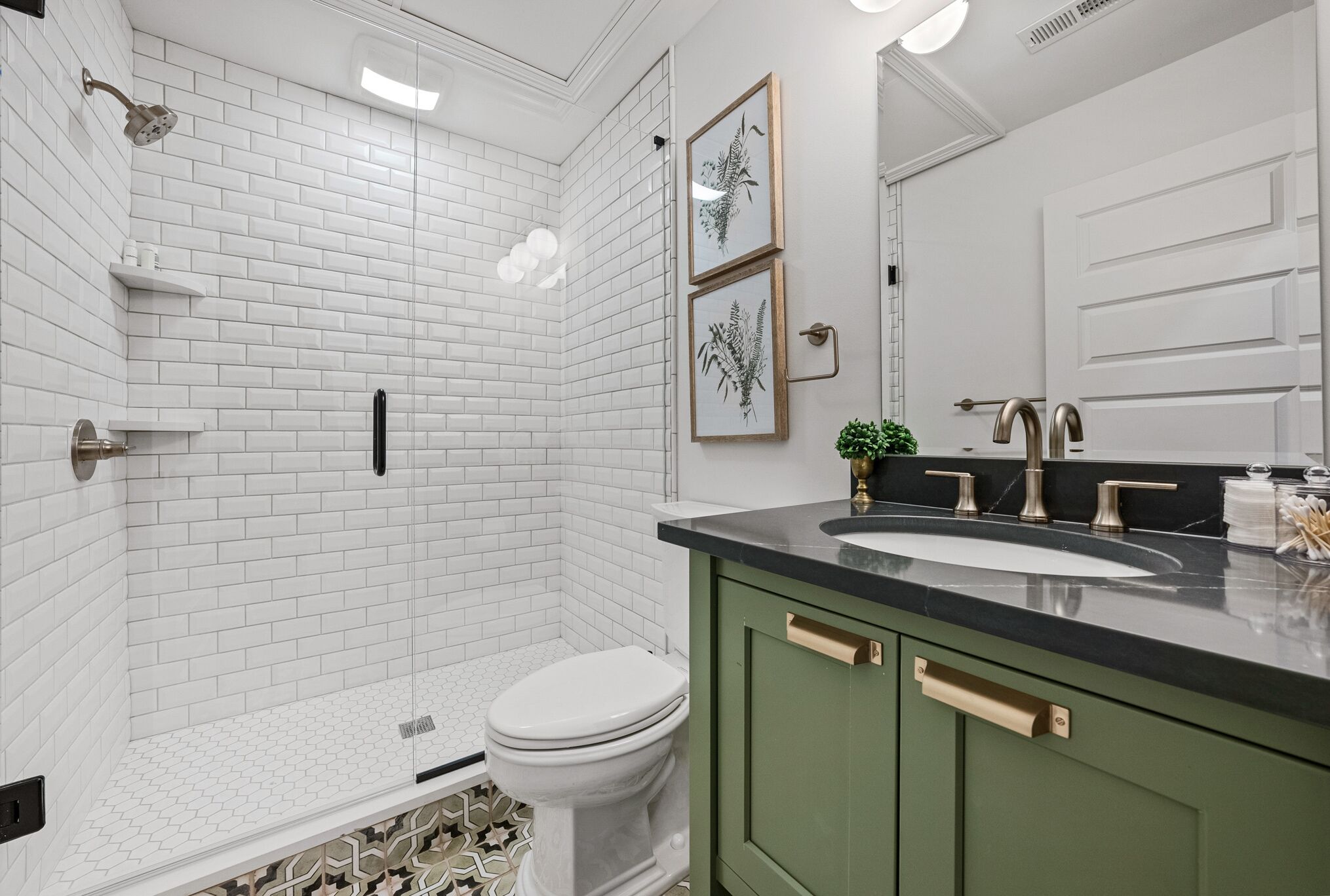 A modern bathroom with white subway tiles, a glass-enclosed shower, a toilet, a sink with a black countertop, and green cabinets.