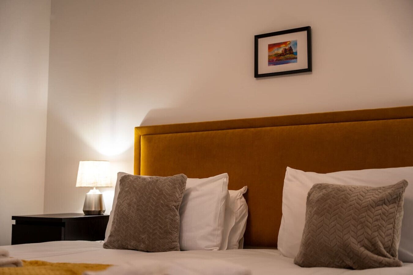 A hotel room with a bed, pillows, a nightstand, a lamp, and a framed picture on the wall.