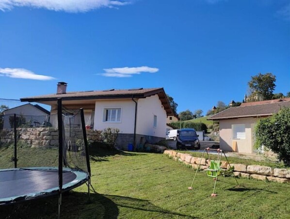 Un quartier résidentiel avec un trampoline au premier plan, une maison avec un mur en pierre et une voiture garée à l’arrière-plan.