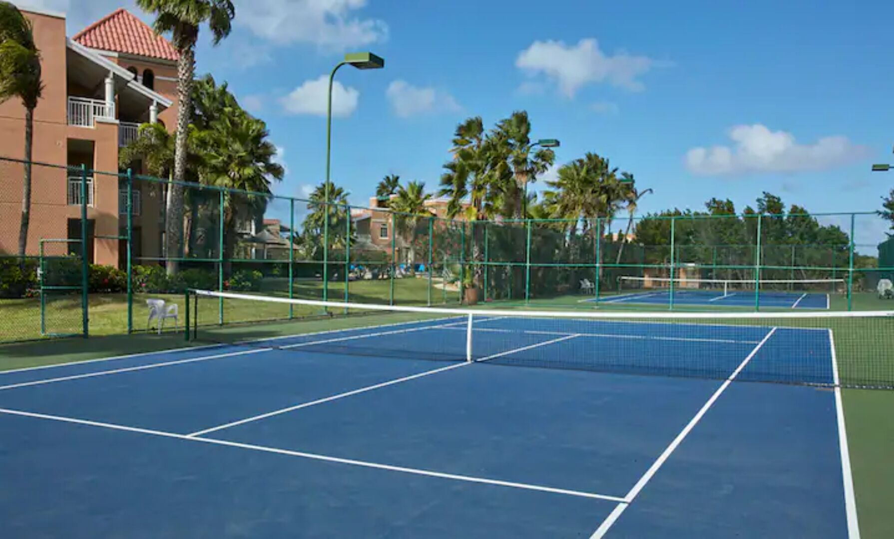 Un court de tennis en plein air, doté d’un filet, entouré d’une clôture et bordé de palmiers.