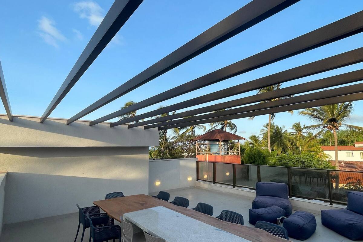 Un patio al aire libre con una mesa larga y sillas, una pérgola de metal, y una vista de palmeras y edificios al fondo.