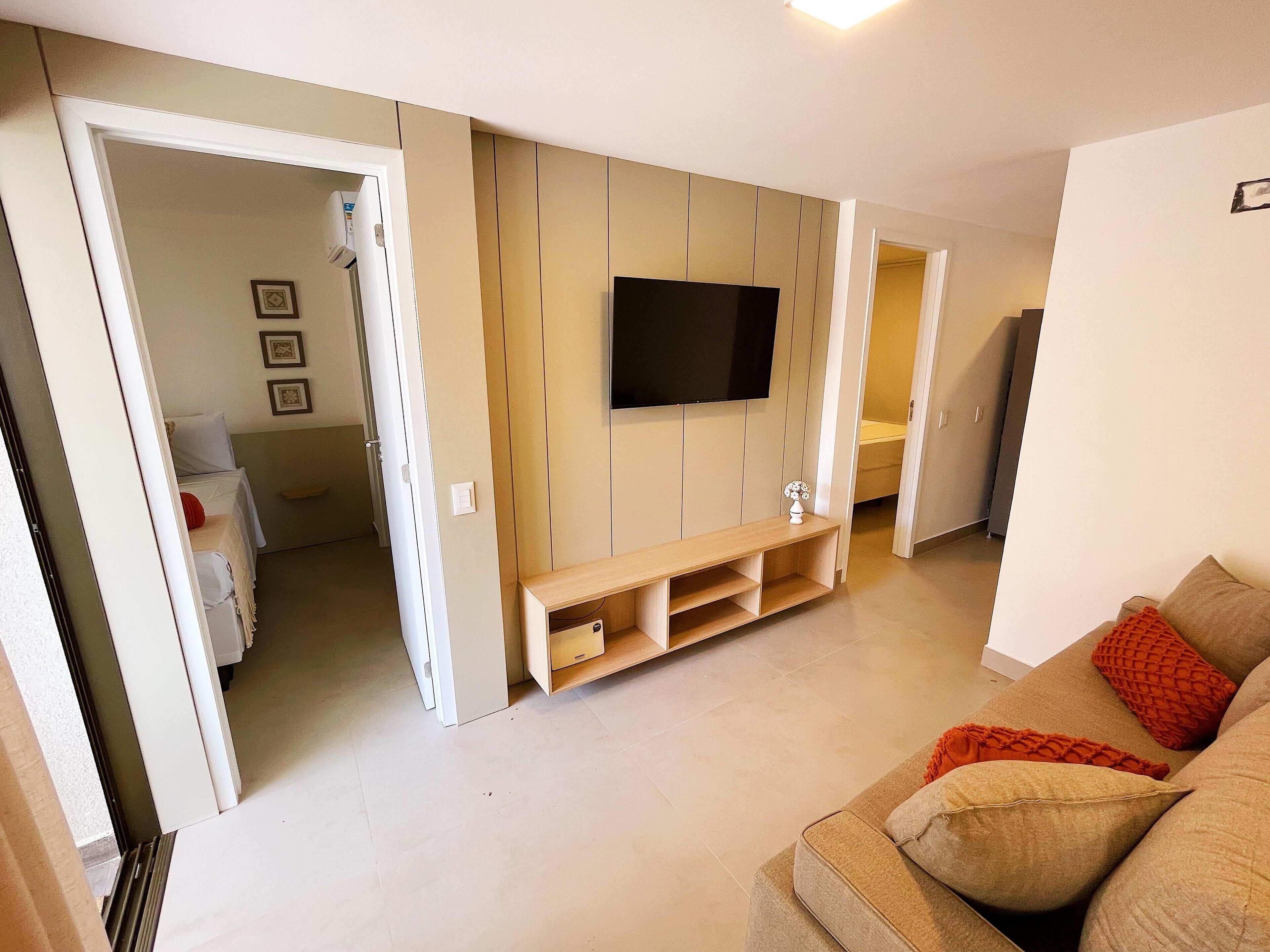 A modern living room with a flat-screen TV mounted on a wooden wall, a wooden entertainment unit, and a beige sofa with orange cushions.