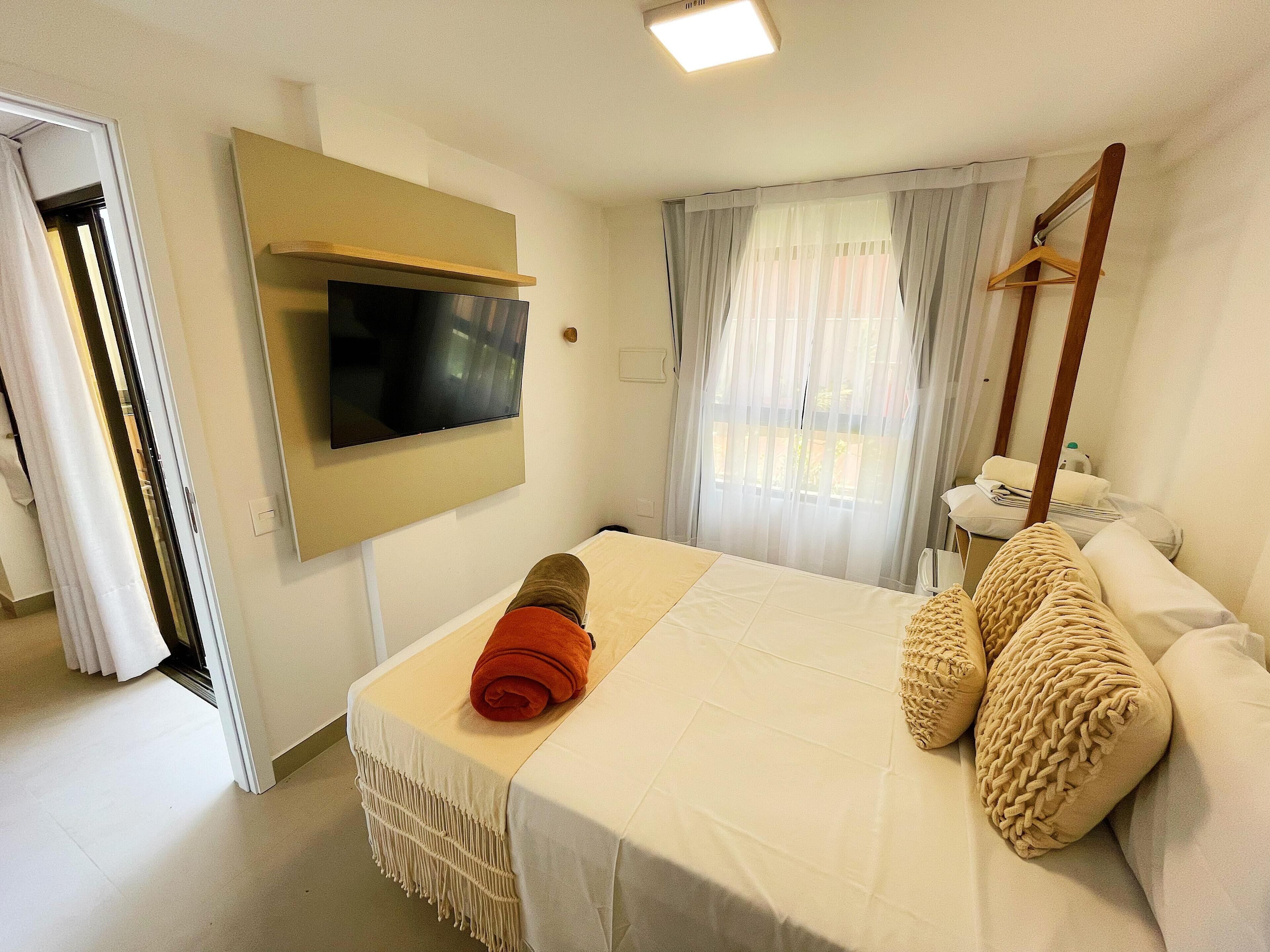 Quarto de hotel com cama, televisão fixada na parede, janela com cortinas e um cabide de madeira na parede.
