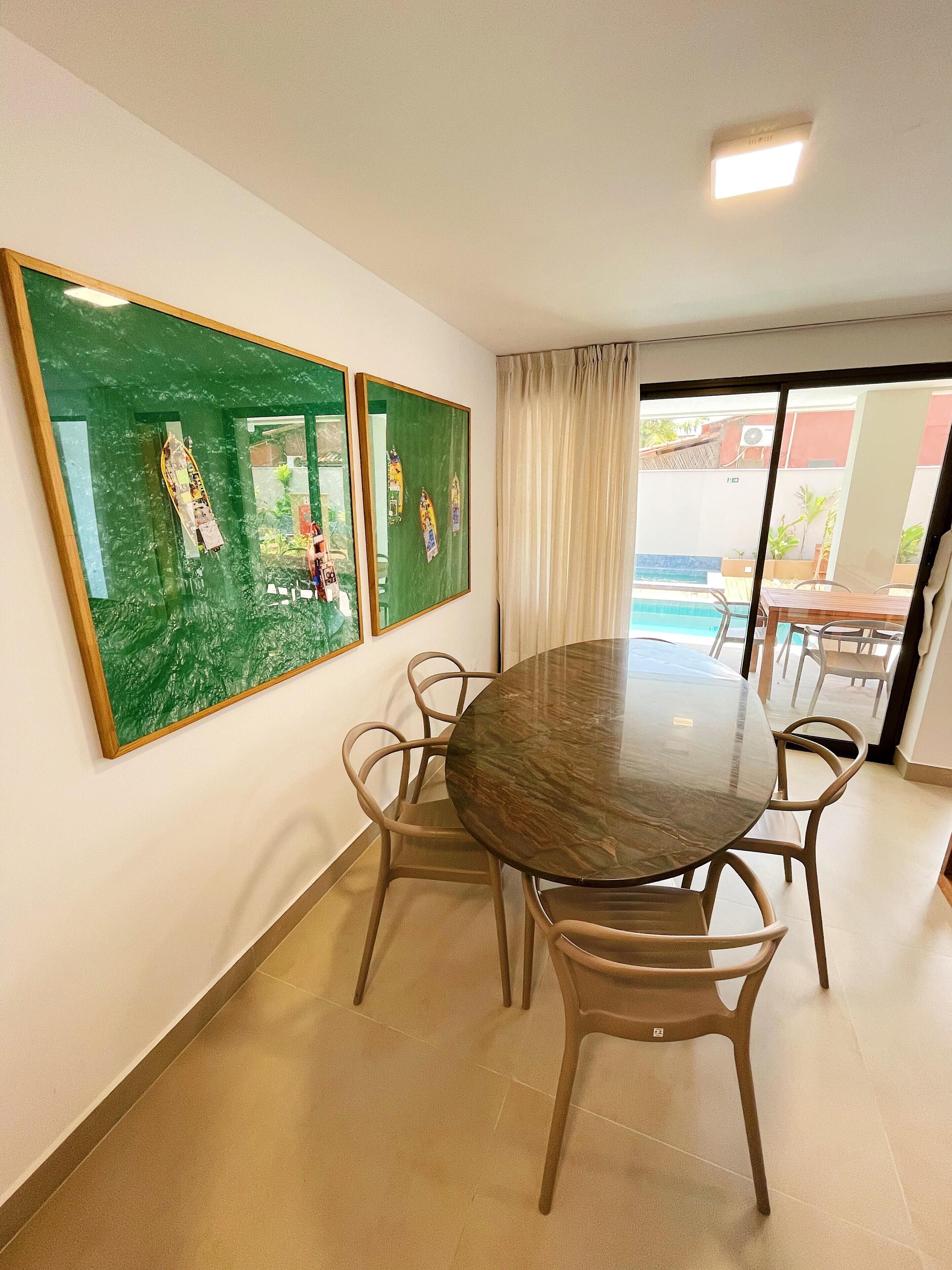 A dining area with a round table and four chairs, a green wall with framed artwork, and a view of a pool through a sliding glass door.