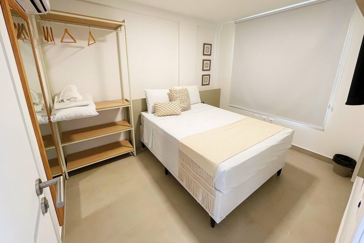 Un dormitorio con una cama, un armario con ropa colgada, una mesita de noche y una ventana.