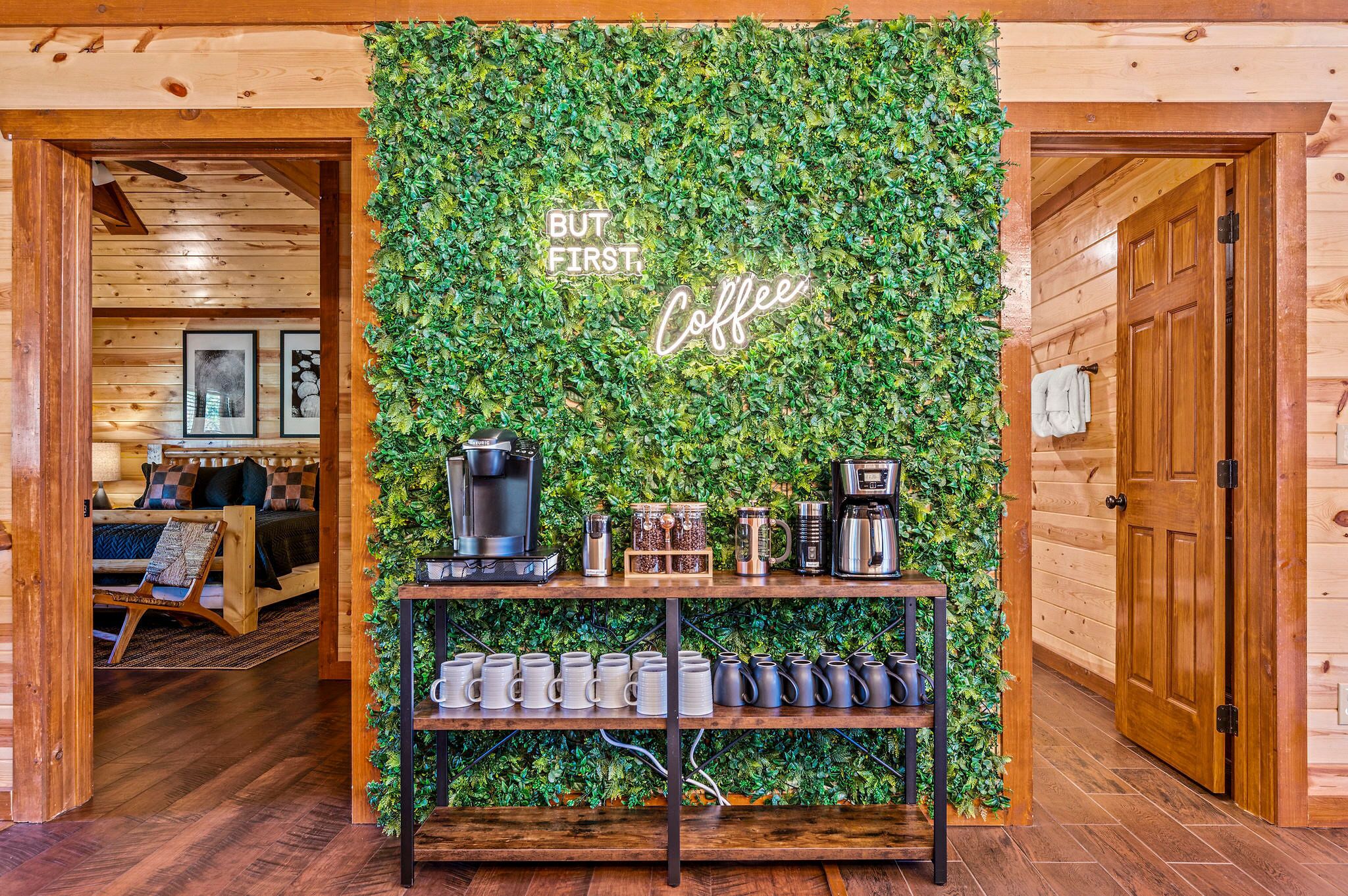 A cozy room with a wooden ceiling, a bed with a checkered blanket, a coffee station with mugs and a coffee maker, and a wall with a green leafy backdrop and a 'But first, Coffee' sign.