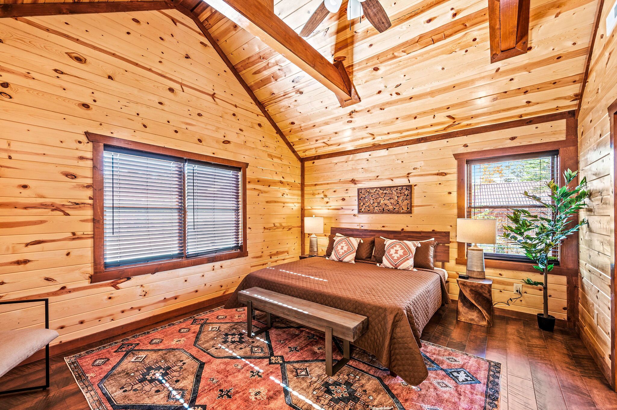 A cozy log cabin bedroom with a large bed, a bench, a rug, and a window with blinds.