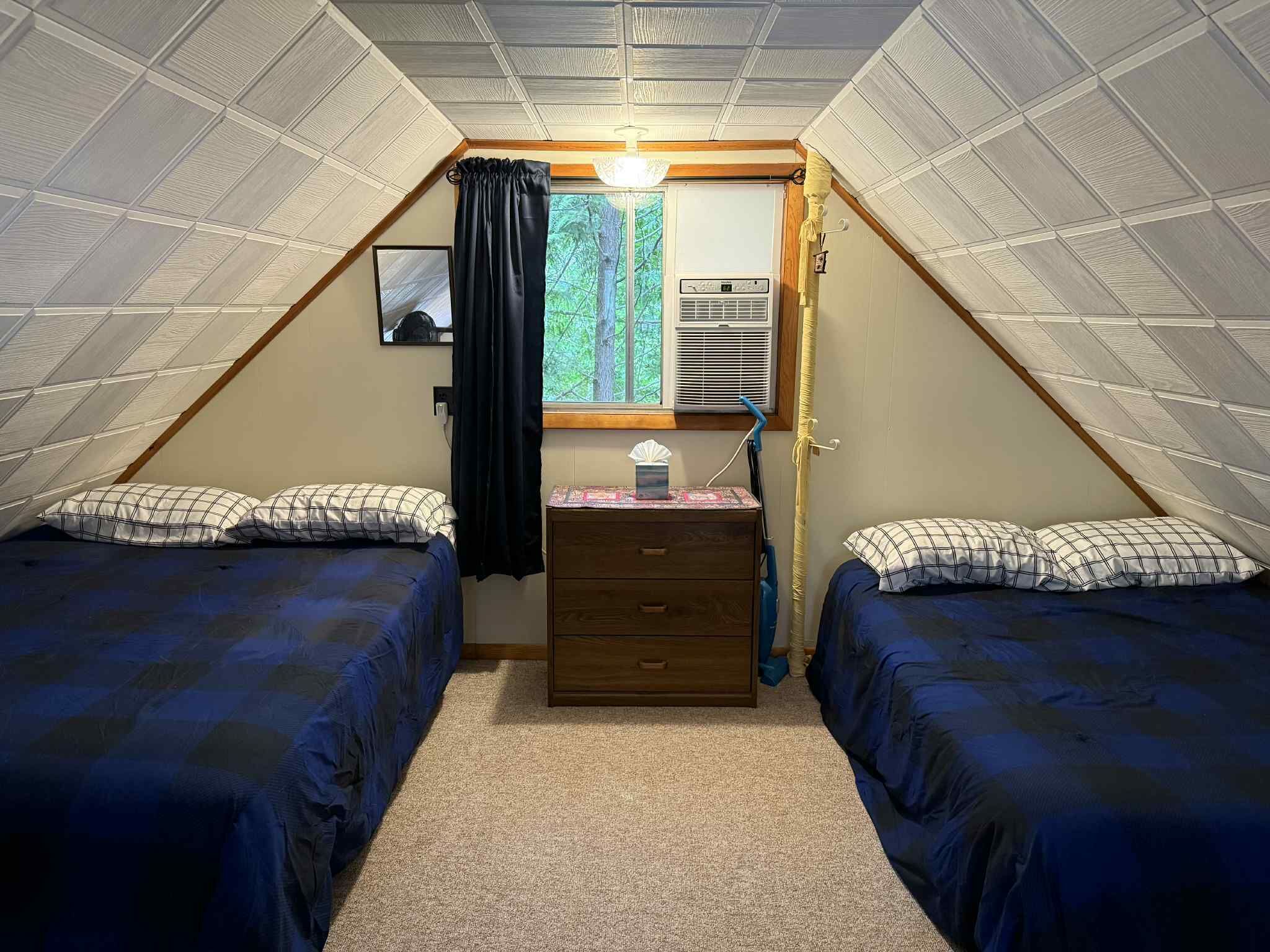 A small, triangular attic bedroom with two beds, a dresser, a window with curtains, and an air conditioning unit.
