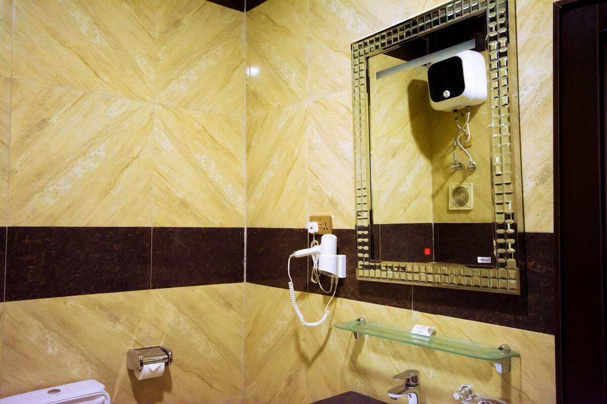 A bathroom with a marble wall, a hairdryer mounted on the wall, a glass shelf, and a toilet.
