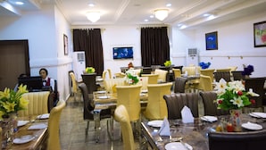 Een restaurantgedeelte van een hotel, met meerdere tafels, stoelen en een televisiescherm.