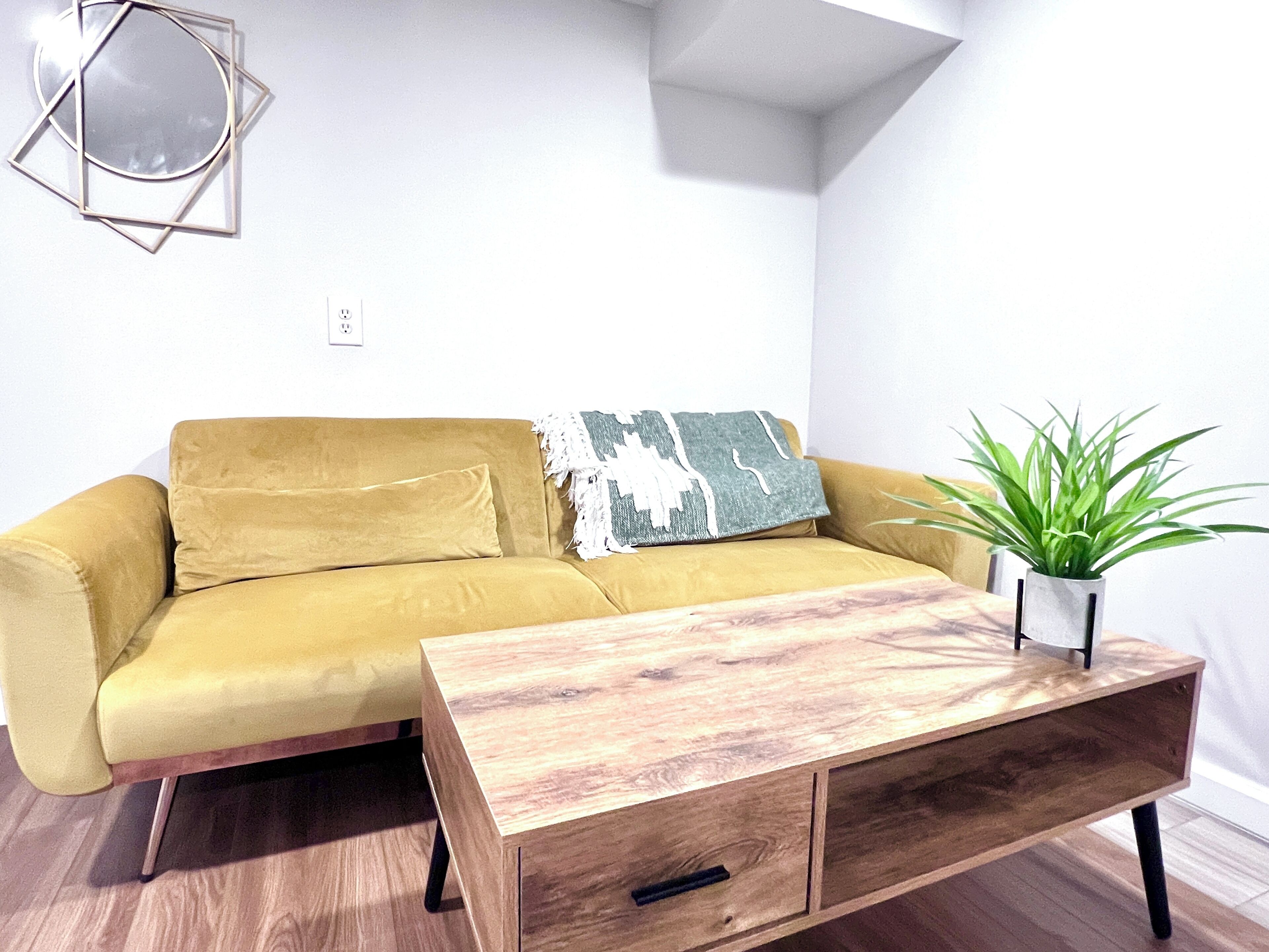Ein senfgelbes Sofa mit einem dekorativen Kissen in geometrischem Muster, ein Couchtisch aus Holz mit Schubladen und eine grüne Pflanze im weißen Topf.