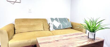 Ein senfgelbes Sofa mit einem dekorativen Kissen in geometrischem Muster, ein Couchtisch aus Holz mit Schubladen und eine grüne Pflanze im weißen Topf.