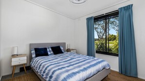 A bedroom with a bed, nightstands, a window with curtains, and a view of greenery outside. - Inverloch' s Lime Green Door - Linen, Wifi, Pet Friendly (Inverloch)