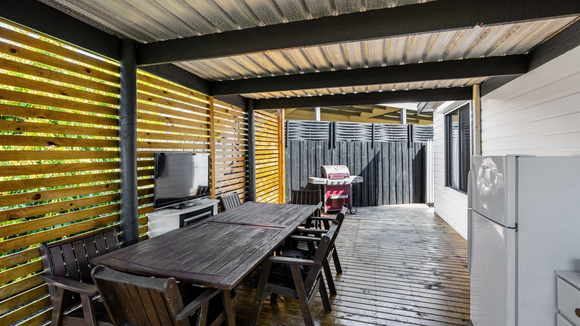 Un espace extérieur abrité, aménagé pour les repas, avec une table et des chaises en bois, un barbecue et un téléviseur.