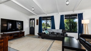 A living room with a flat-screen TV, a leather sofa, a coffee table, and a large window with curtains. - Inverloch' s Lime Green Door - Linen, Wifi, Pet Friendly (Inverloch)