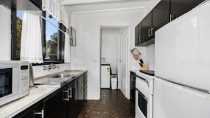A narrow kitchen with white cabinets, a microwave, and a stove. There is a window with lace curtains and a view of trees outside. - Inverloch' s Lime Green Door - Linen, Wifi, Pet Friendly (Inverloch)