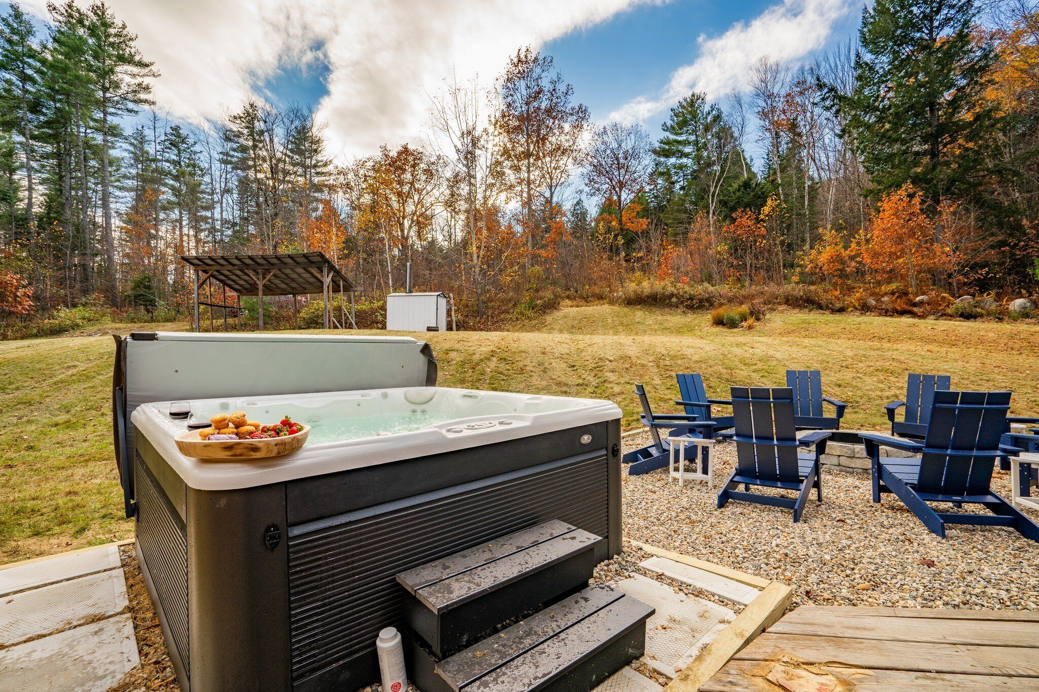 A hot tub with steps and a wooden deck, surrounded by Adirondack chairs and a forest backdrop.
