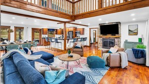 A spacious living area with a high ceiling, wooden beams, a fireplace, and a dining area with a view of the outdoors. - Sleeps 20 | Hot Tub | Game Room | Fire Pit |4500SF (Newry)