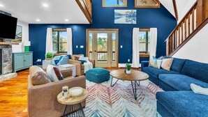 A modern living room with a blue accent wall, a wooden staircase, and a large window with a view of trees. - Sleeps 20 | Hot Tub | Game Room | Fire Pit |4500SF (Newry)