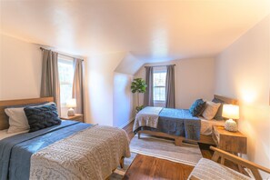 Un dormitorio acogedor con dos camas, una mesita de noche con lámpara, una ventana con cortinas y una planta.