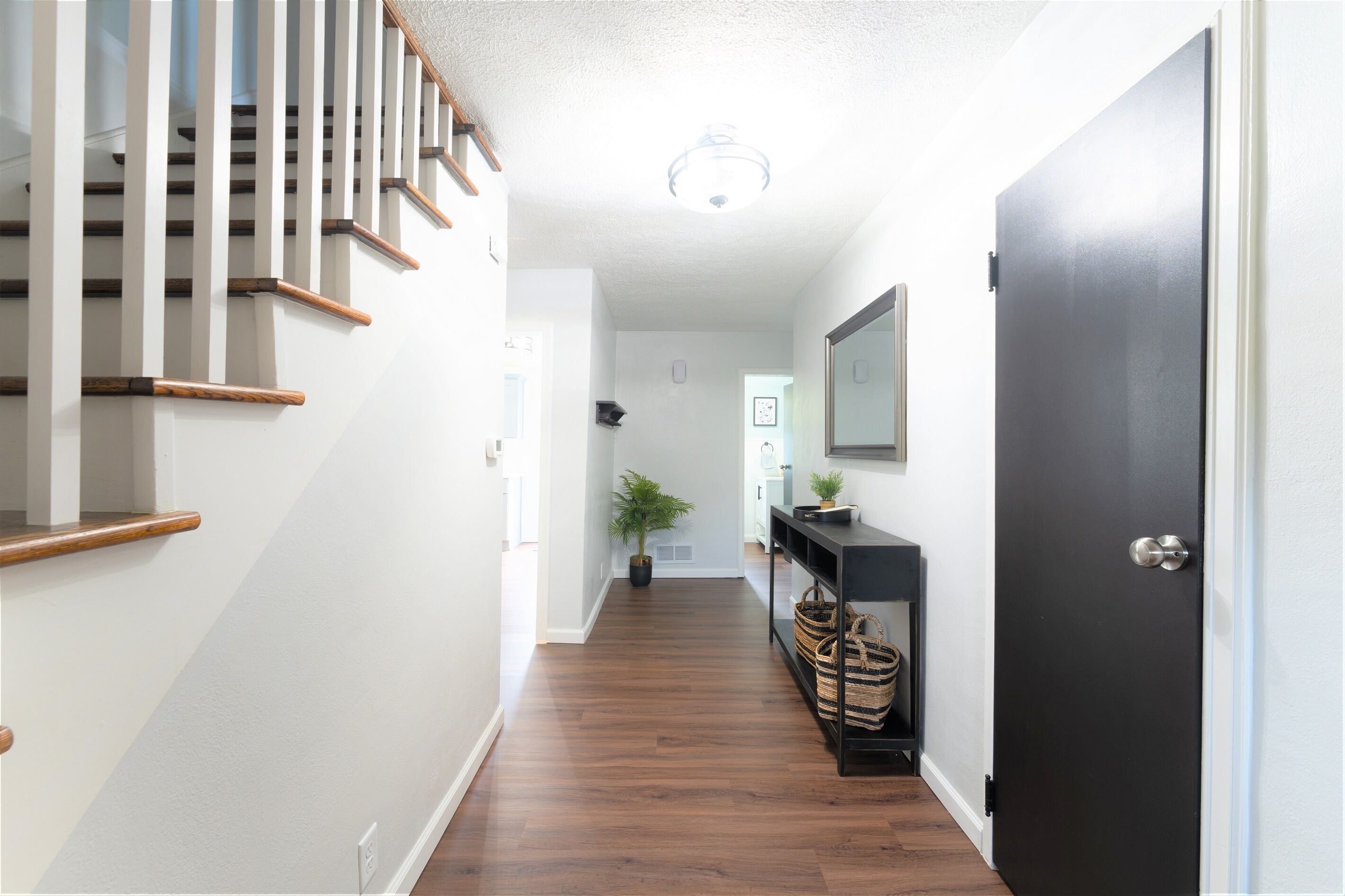 Un couloir avec un escalier en bois, une porte noire, une plante en pot et une console surmontée de paniers.