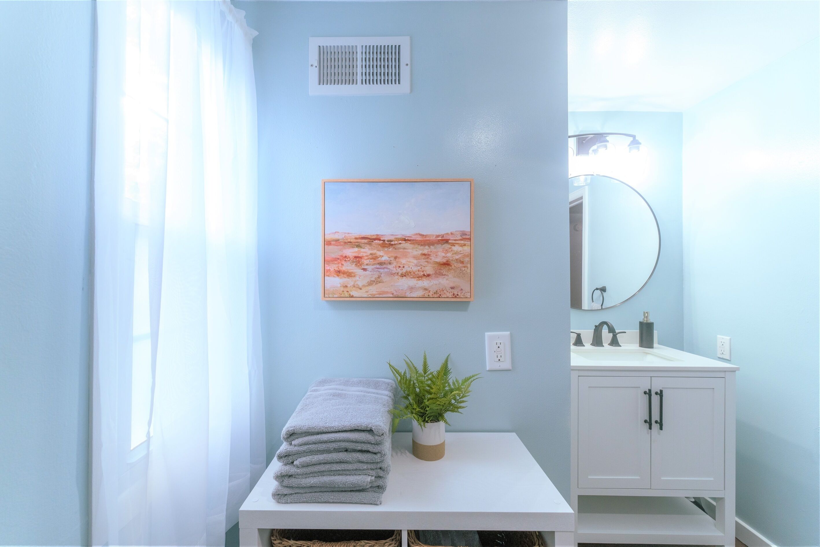 Une salle de bain avec un meuble-lavabo blanc, un évier, un miroir, un tableau encadré, une plante et une pile de serviettes.