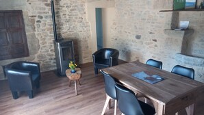 Een rustieke kamer met een stenen muur, een houten tafel met stoelen, een klein houten krukje met een vaas en een zwarte verwarming.